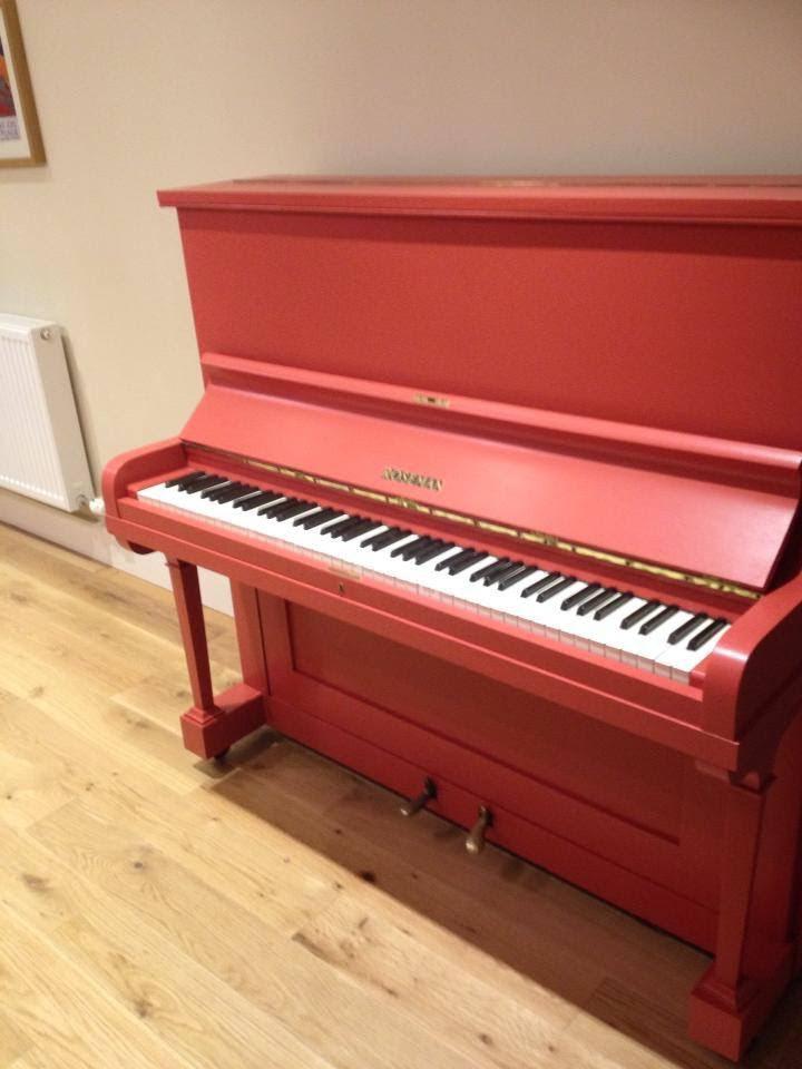 Red upright piano placed against a painted interior wall on wooden flooring.