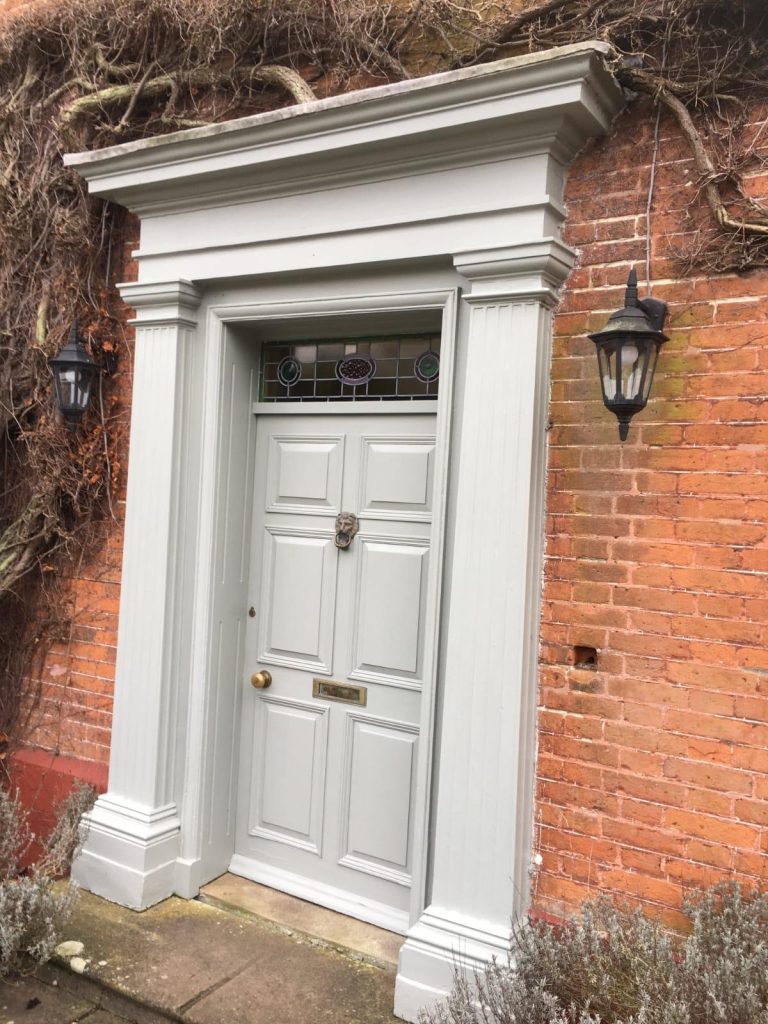 Grey painted front door with decorative surround set within a red brick wall.