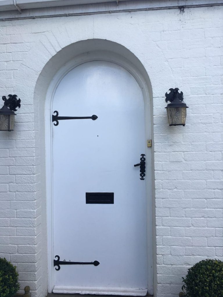 White arched front door with black iron hinges set in a painted brick wall.