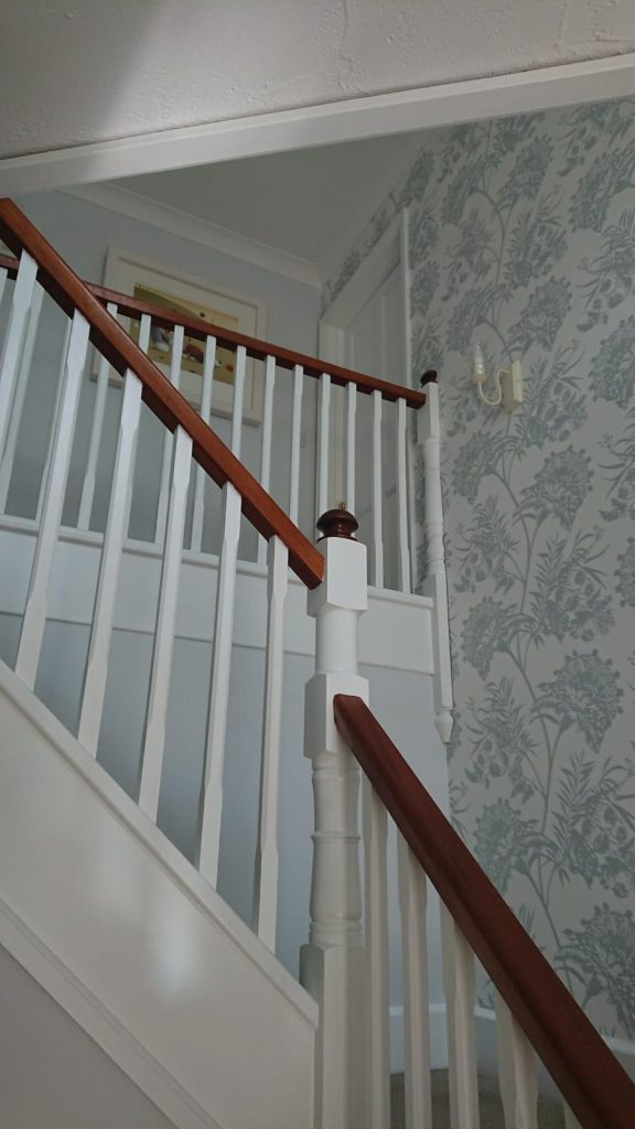 White painted staircase with wooden handrail and floral patterned wallpaper.